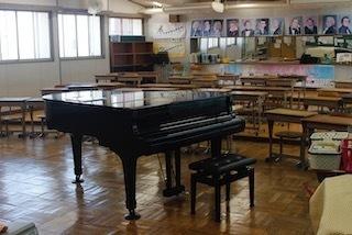 前方にグラウンドピアノが置かれ、段上がりに生徒の机と椅子が設置され、後ろの壁面には有名な世界の作曲家の肖像画が掲示されている音楽室の写真